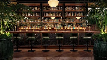 A bar with multiple shelves of liquor bottles, green bar stools, and large potted plants.