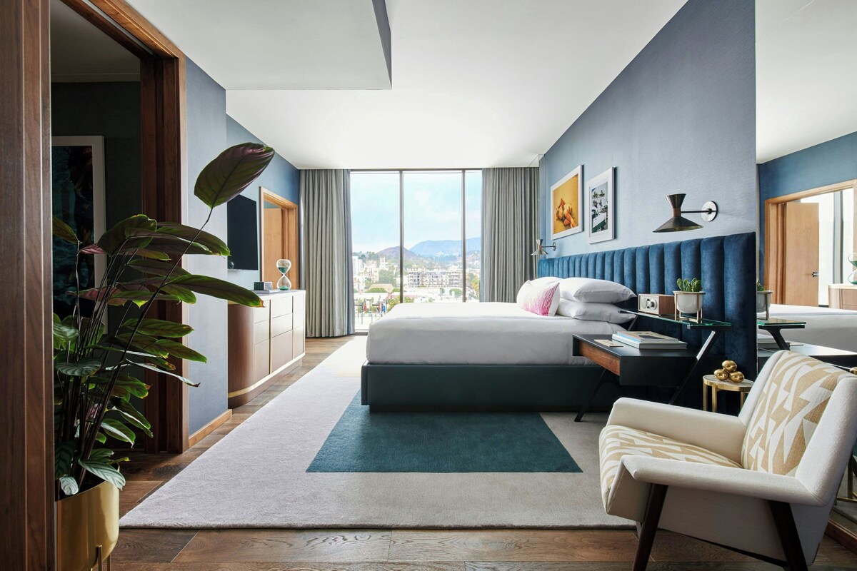 A modern hotel room with a large bed, a desk, a chair, and a view of the city.
