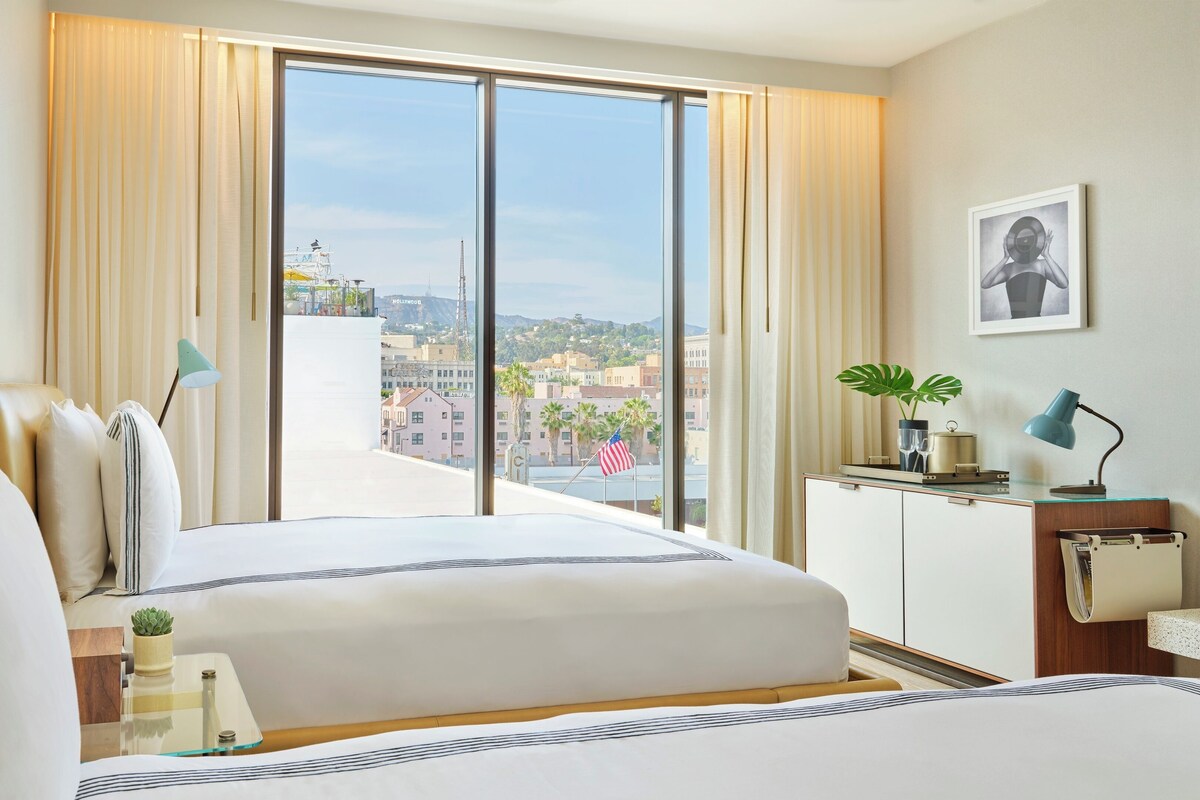A hotel room with a large window, a bed, a desk, and a view of a cityscape.