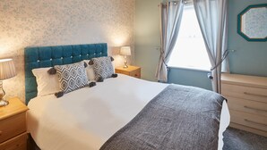 A neatly made bed with a blue headboard, a bedside table with a lamp, and a dresser with drawers. - Apartment 4 At Glencoe (Whitby)