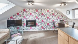 A loft-style living space with a sloped ceiling, a flat-screen TV, a glass coffee table, a wicker basket, a dining area with a wooden table and chairs, and a floral-patterned wall. - Apartment 4 At Glencoe (Whitby)