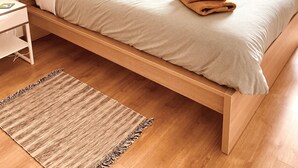 Ein ordentlich gemachtes Bett mit Kissen und einem gefalteten Handtuch, ein Nachttisch mit Lampe, ein gerahmtes Bild an der Wand und ein Fenster mit Vorhängen.