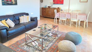 Ein Wohnzimmer mit schwarzem Ledersofa, einem Glastisch, einem Esstisch mit Stühlen und einem bunten Teppich.