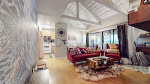Ein geräumiges Wohnzimmer mit einem roten Sofa, einem Holztisch und einem Fellteppich. Im Hintergrund ist eine Küche mit modernen Geräten zu sehen.