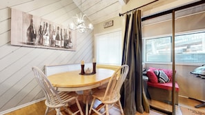 A dining area with a round table and four chairs, a chandelier, a wall-mounted artwork of wine bottles, and a sliding glass door leading to a balcony with a sofa and a grill. - Rustic Mountain Cabin, Outdoor Pool & Hot Tub On-site. Golf Course Nearby! (Mammoth Lakes)