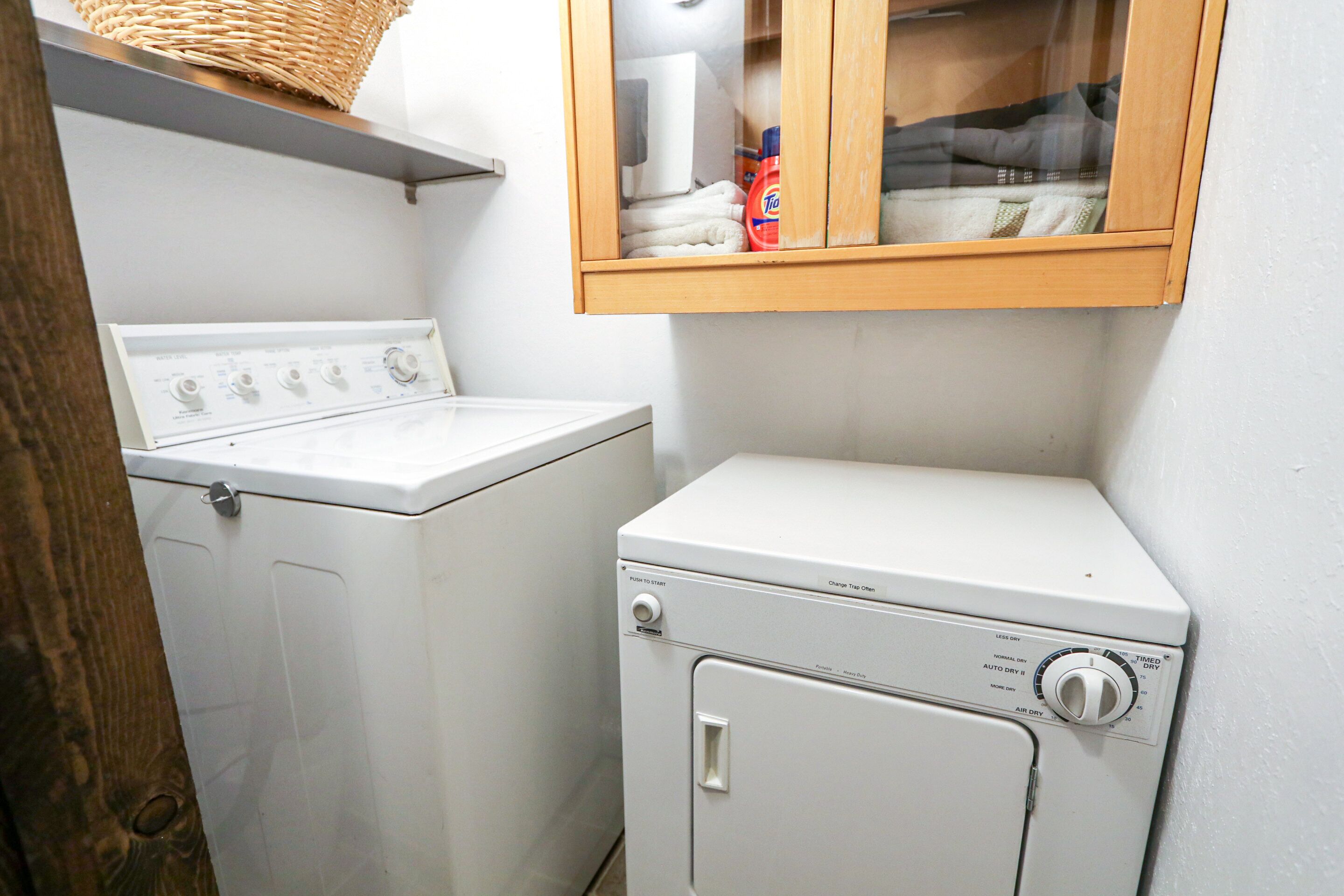 A laundry area with a washing machine and dryer, a wooden cabinet above, and a shelf with laundry supplies.