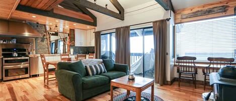 A spacious living area with a green sofa, wooden flooring, a dining table with chairs, a kitchen with modern appliances, and a large window with curtains.
