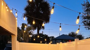 An outdoor seating area with Adirondack chairs and a fire pit, surrounded by a white fence and string lights. - Beautiful home with pool. See notes about heated pool season. (Tybee Island)
