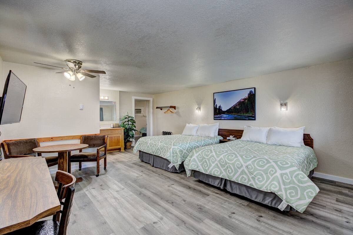 A hotel room with two beds, a desk, chairs, a ceiling fan, and a painting on the wall.