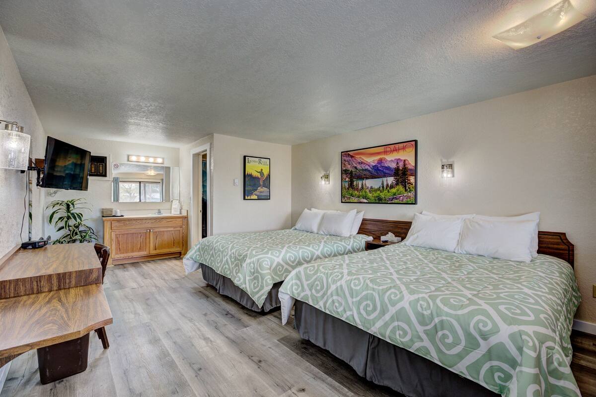 A hotel room with two beds, a wooden desk, a small kitchenette, and a bathroom with a shower.