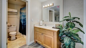 A bathroom with a wooden vanity, a sink, a mirror with lights, a toilet, and a shower curtain. - Weiser Motel & RV Park (Weiser)