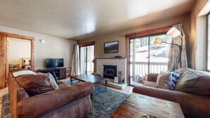 A cozy living room with a fireplace, comfortable seating, a television, and a large window with a view of the outdoors. - Quiet Forest Setting. Near The Village & Lake Basin, Pool & Hot Tub On-site (Mammoth Lakes)