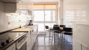 A modern kitchen with white cabinetry, a large window with blinds, and a dining area with black chairs. - FLH Cascais Wooden Spacious Apartment (Cascais)