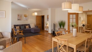 A modern living room with a wooden dining table, sofas, and a coffee table. There are wall-mounted lights and a door leading to another room. - FLH Cascais Wooden Spacious Apartment (Cascais)