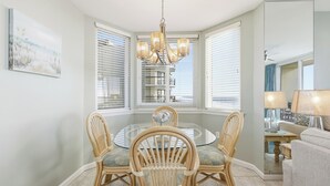 A dining area with a round glass table, four wicker chairs, a chandelier, and a view through large windows with blinds.