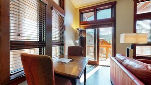 A room with a wooden table, a cushioned chair, a sofa, and large windows with blinds. - Modern Home, Great Mountain Views, Private Hot Tub, Gas Grill & Washer/Dryer (Mammoth Lakes)