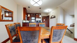 A dining area with a wooden table and chairs, a kitchen in the background, and a staircase leading to another floor.