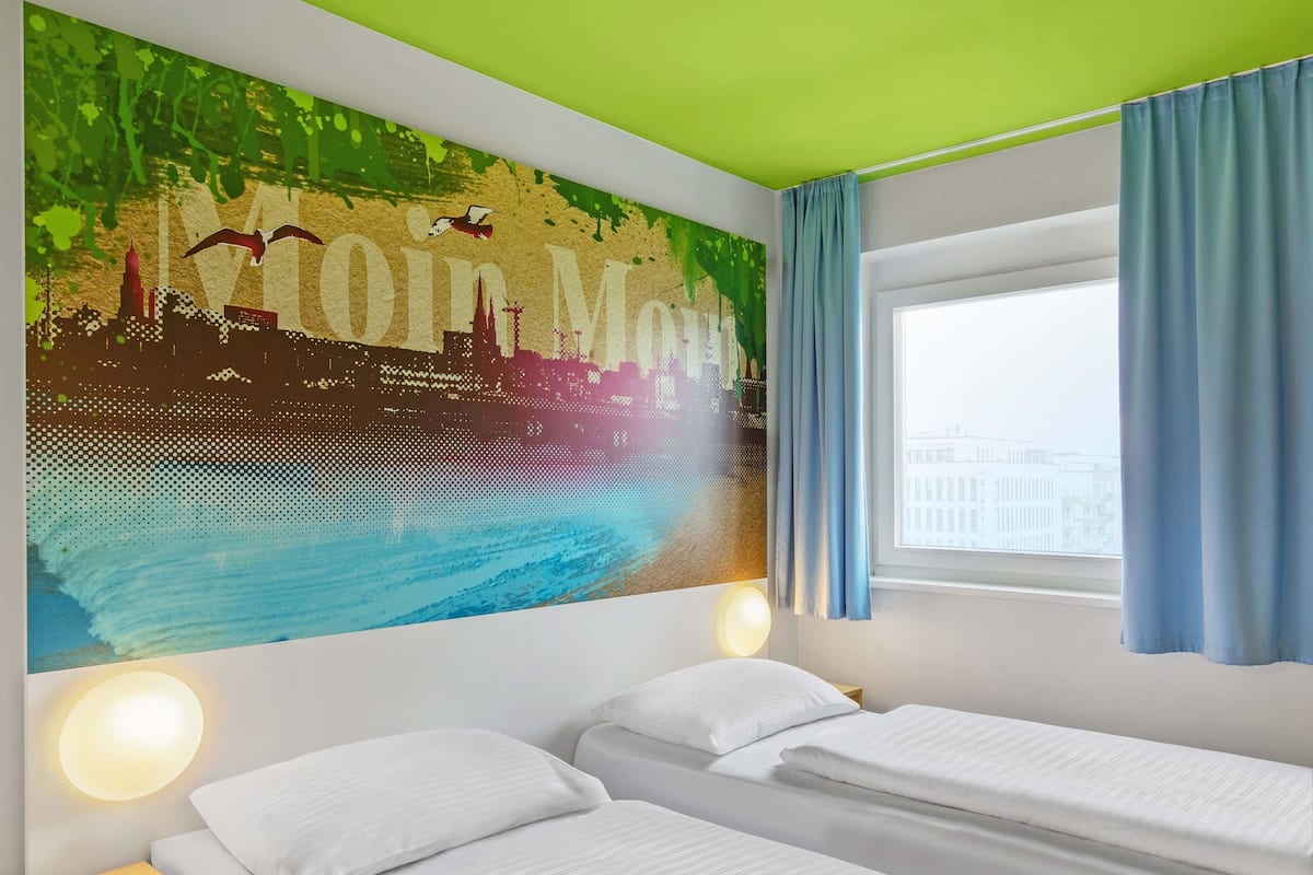 A hotel room with two beds, a cityscape mural, a green ceiling, and a window with curtains.