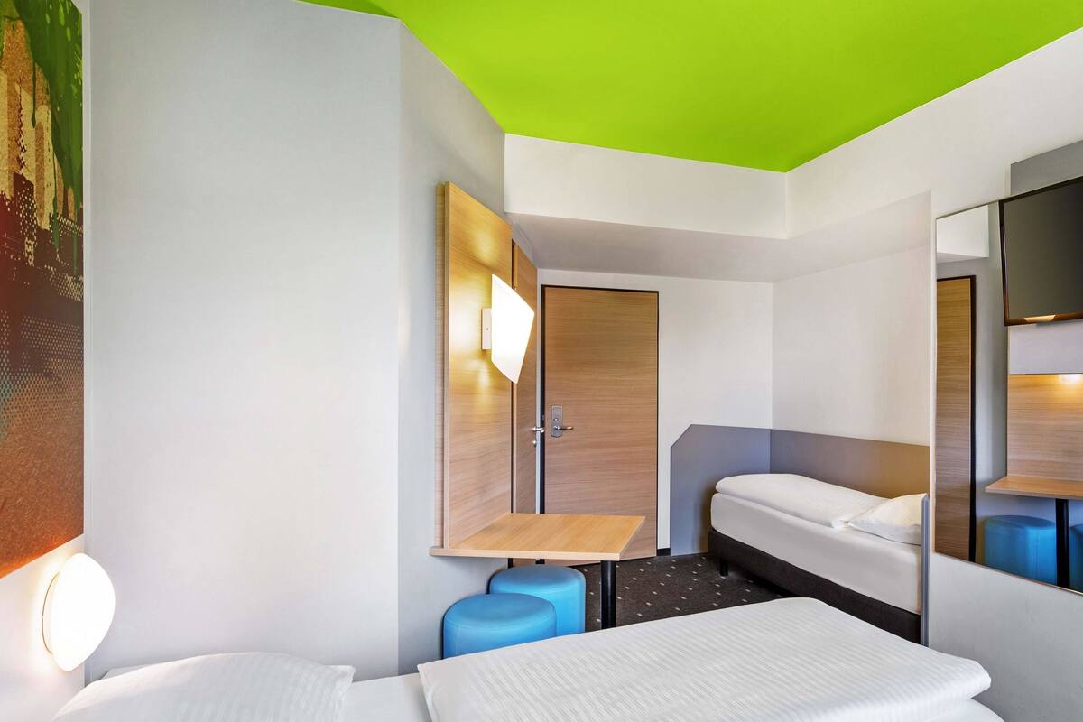 A hotel room with a bed, a desk, a chair, a mirror, and a door.