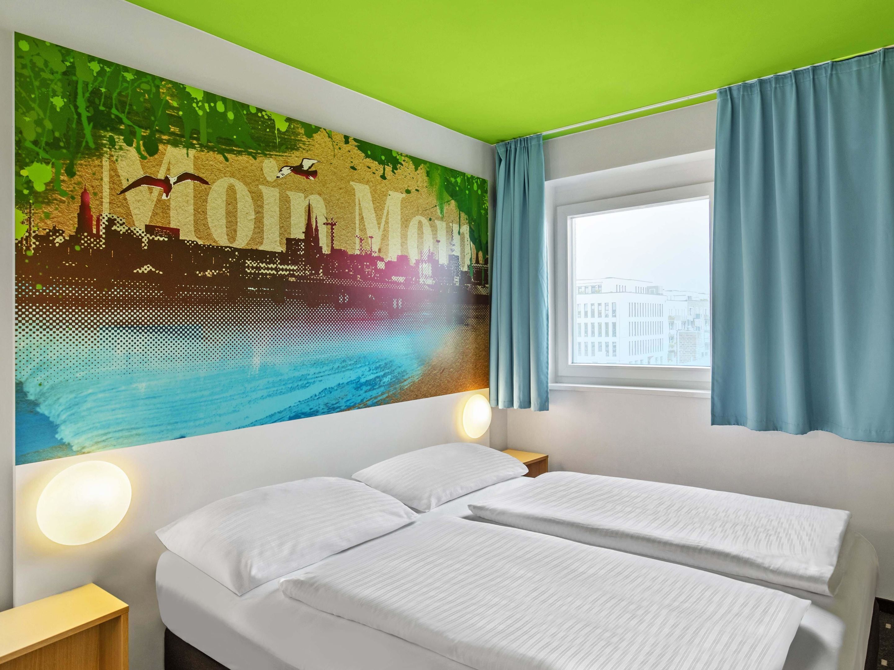 A hotel room with a bed, two bedside tables, a wall mural, a window with curtains, and a green ceiling.