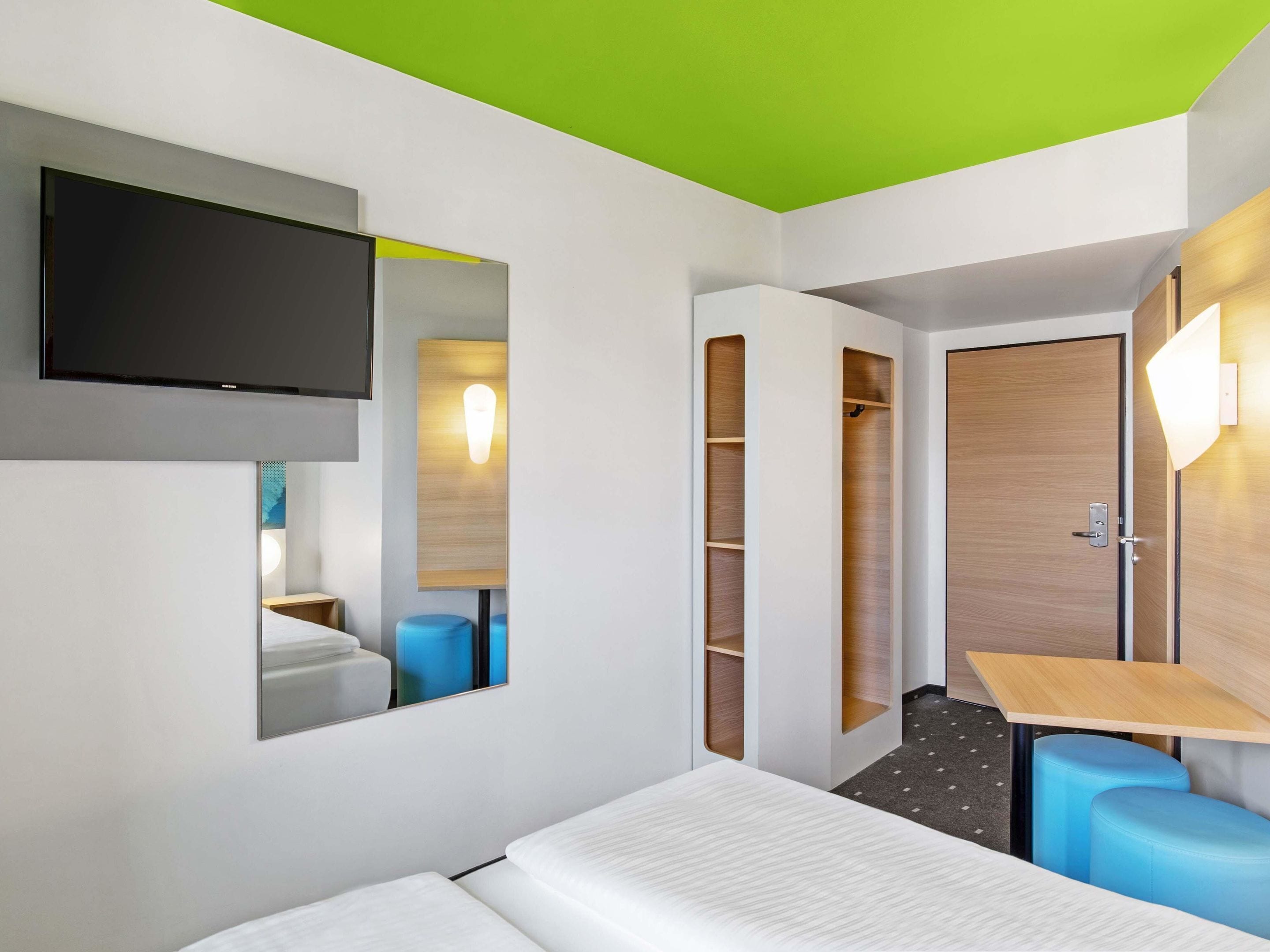 Ein modernes Hotelzimmer mit Bett, Wand-Fernseher, Schrank mit integrierter Beleuchtung und kleinem Tisch mit Stuhl.