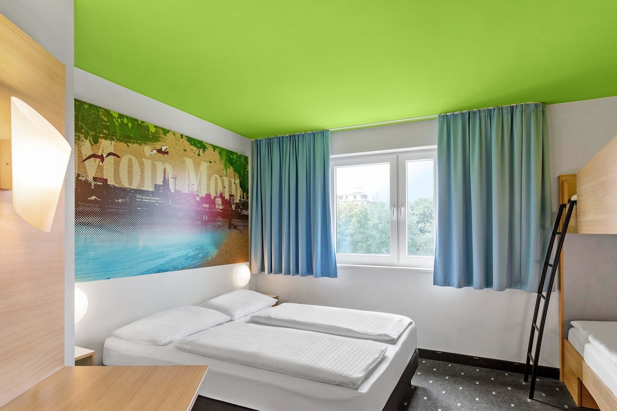 Une chambre d’hôtel avec un lit, une fenêtre avec des rideaux, un mur orné d’une fresque représentant un paysage urbain, et une échelle.