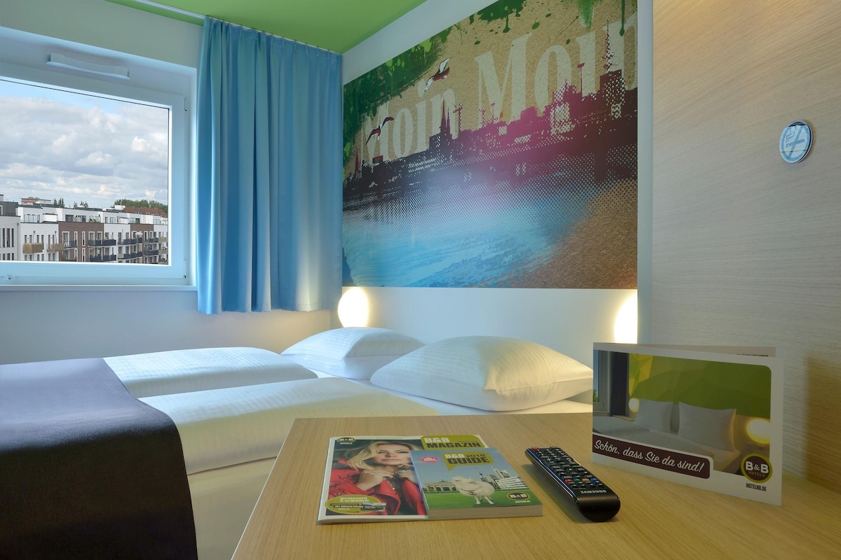 Une chambre d’hôtel avec un lit, une télécommande, un magazine et une vue sur la ville par la fenêtre.