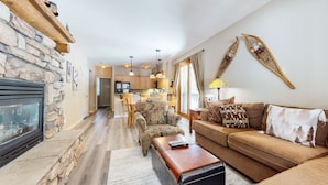 A living room with a stone fireplace, a sofa with patterned cushions, a coffee table, and a kitchen in the background. - On Sierra Star Golf Course, Bright Unit, Private Gas Grill, Pool On-site (Mammoth Lakes)