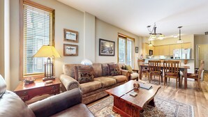 A living room with a sofa, coffee table, and a dining area with chairs and a table. There is a kitchen in the background with cabinets and a refrigerator.