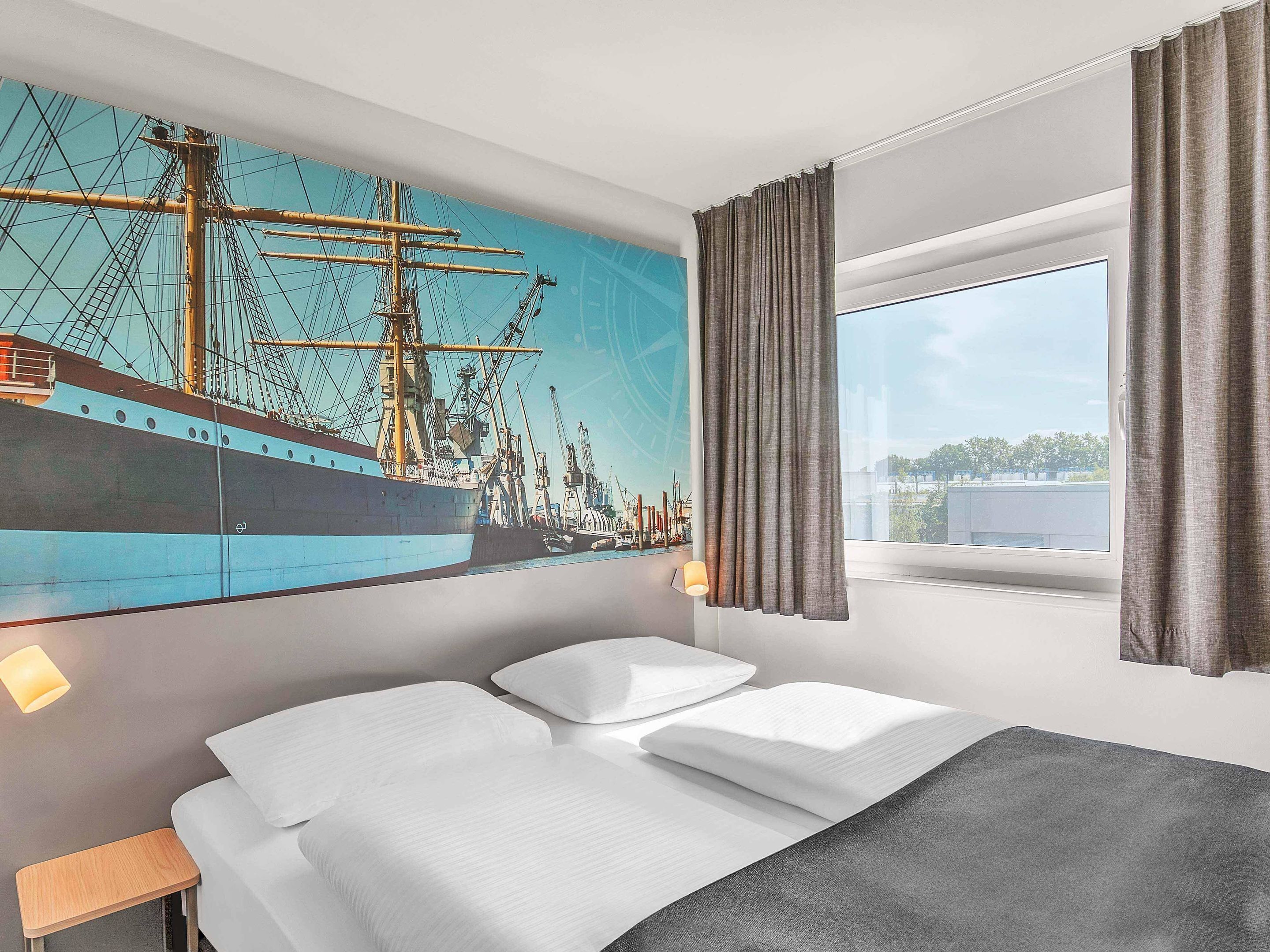 A hotel room with a large bed, a nightstand, a window with curtains, and a wall mural of a ship.