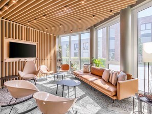 A modern lounge area with a wooden ceiling, a large flat-screen TV, comfortable seating, and large windows offering a view of the city.