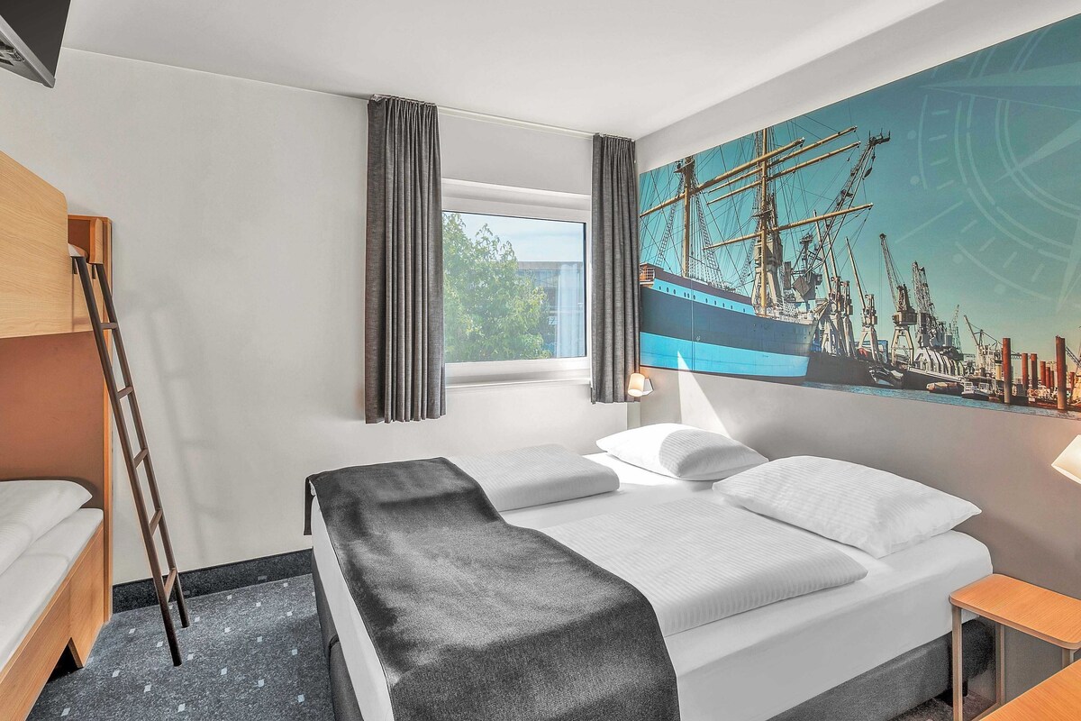 A hotel room with a bed, a desk, a window with curtains, a painting of a ship, and a ladder.