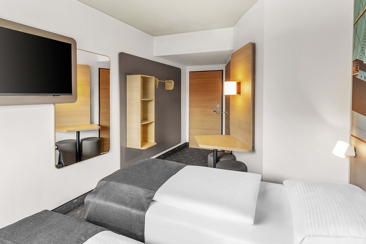 A modern hotel room with a bed, a flat-screen TV, a wooden wardrobe, a mirror, and a wall-mounted lamp.
