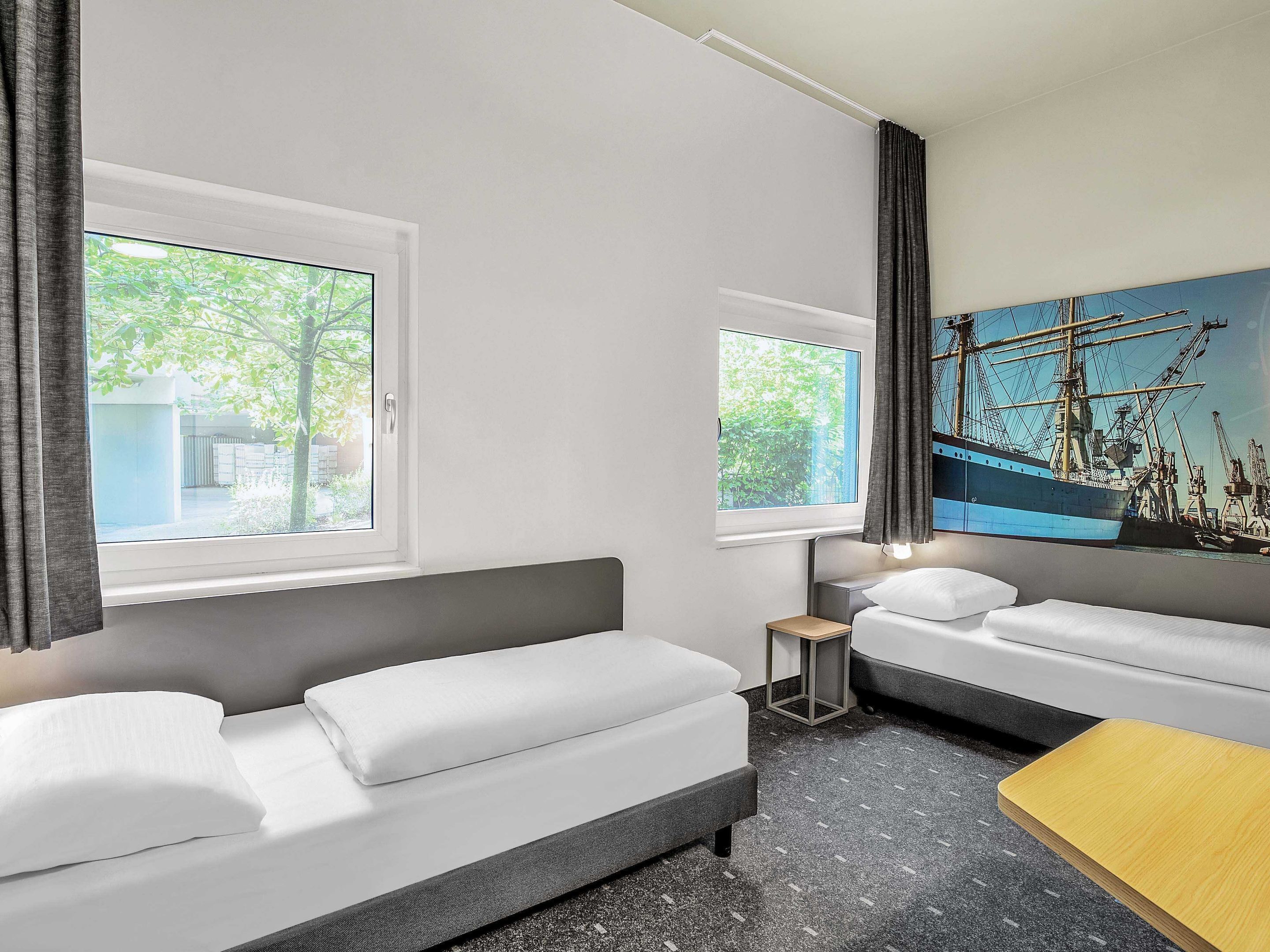 Quarto de hotel moderno com duas camas, uma mesinha e um quadro de um navio na parede.