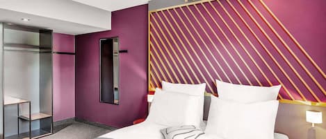 A modern hotel room with a large bed, a bedside table with a lamp, a wardrobe, and a mirror on the wall.