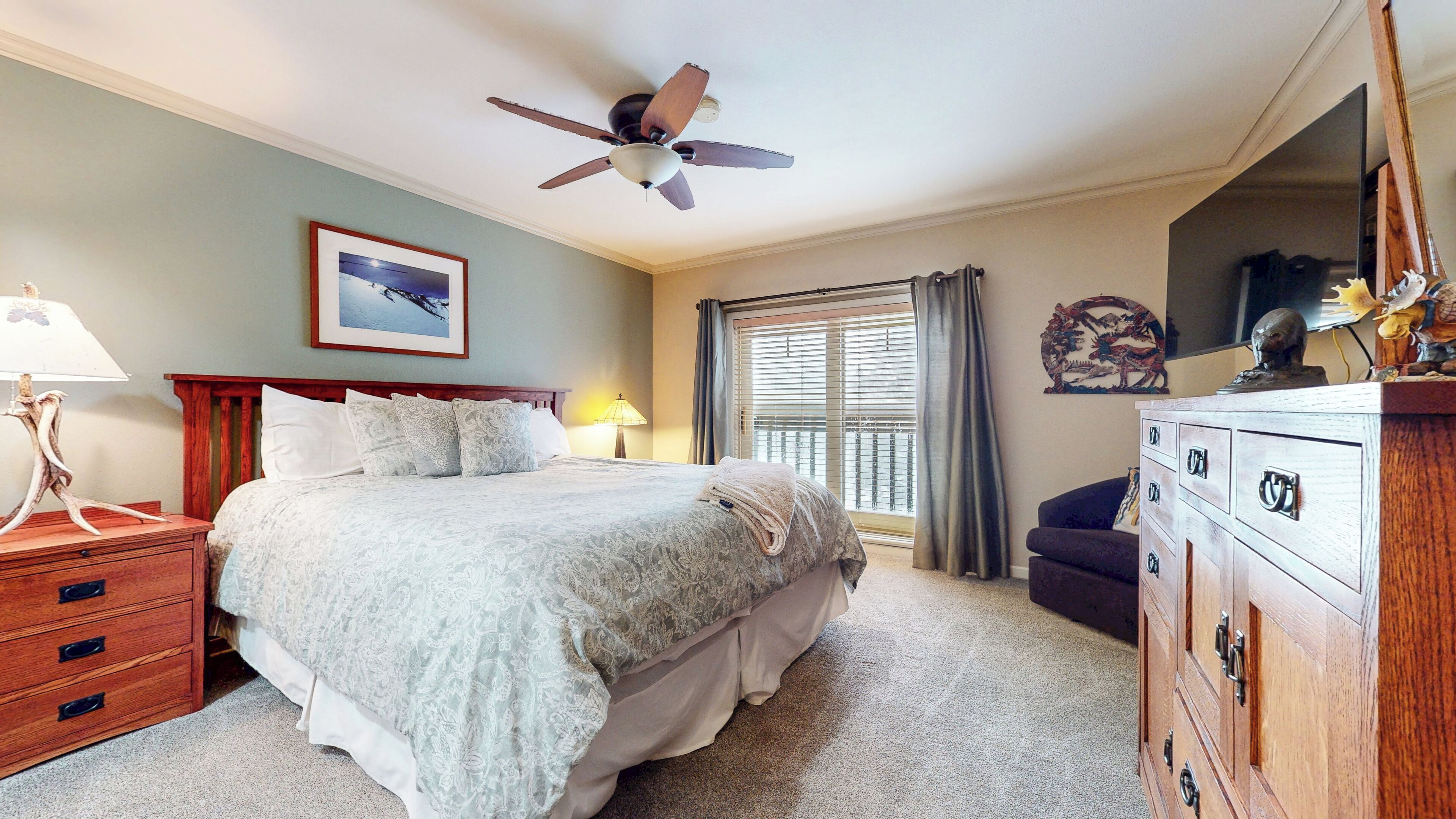 Une chambre à coucher comprenant un lit, une table de chevet, une commode, un ventilateur de plafond, une fenêtre avec des rideaux et un tableau accroché au mur.