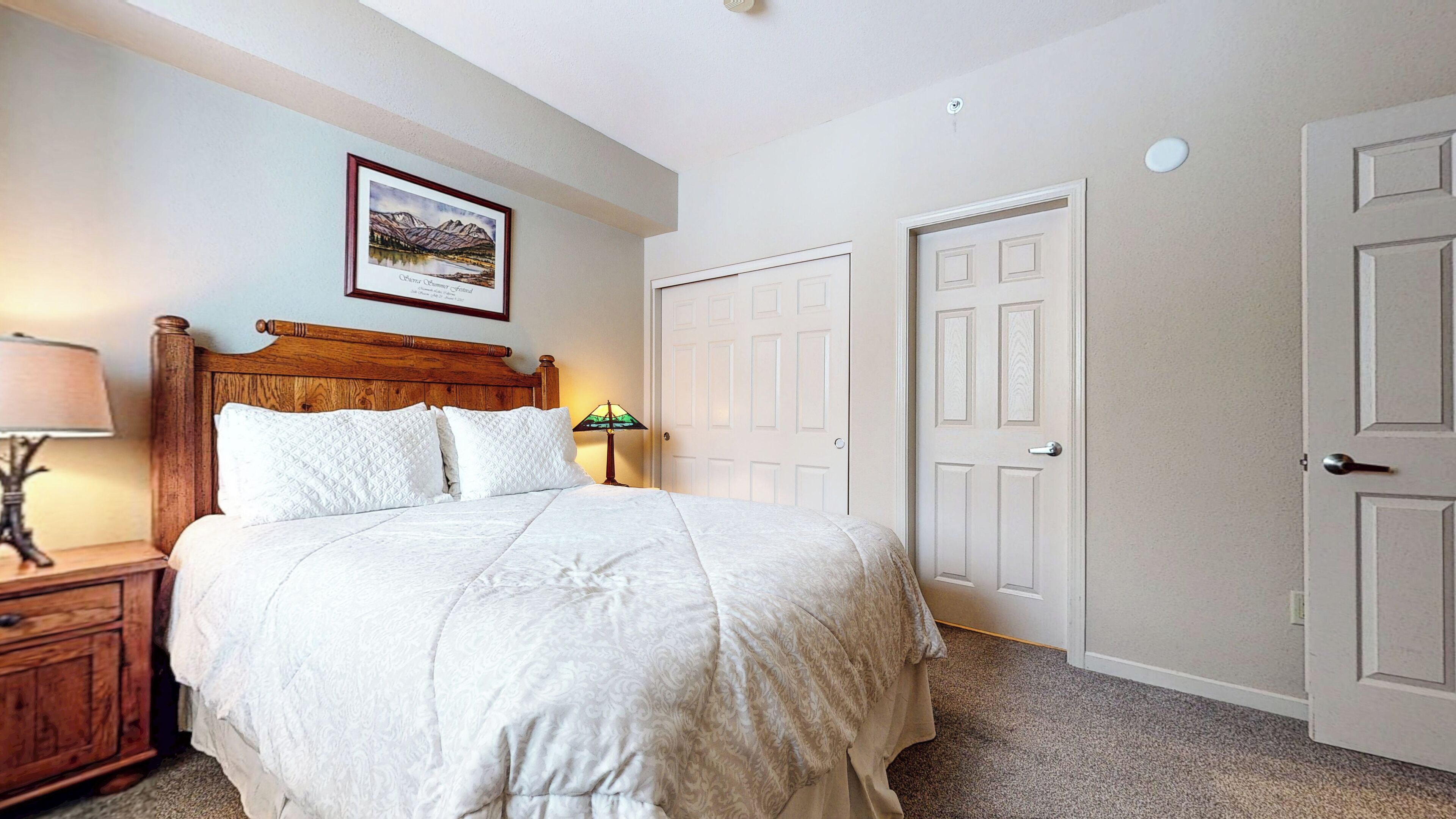Une chambre à coucher bien rangée, avec une tête de lit en bois, un lit recouvert d’une literie blanche, une table de chevet avec une lampe et un tableau encadré accroché au mur.