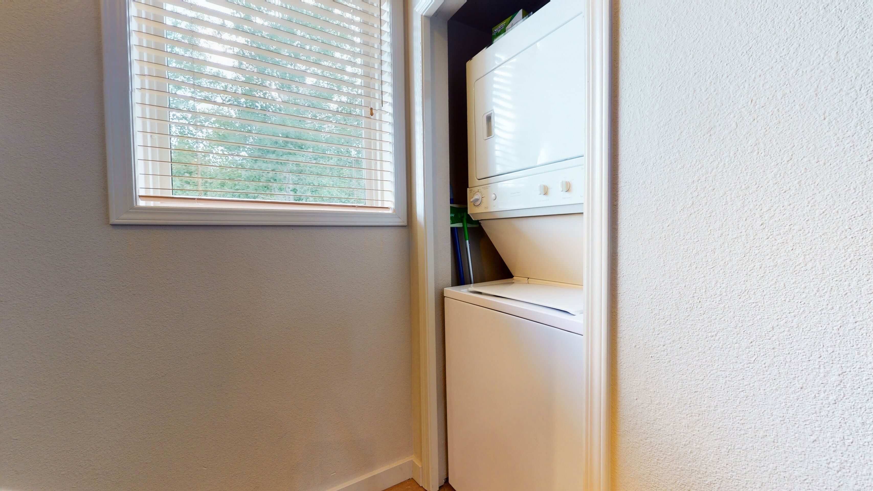 A laundry area with a washing machine and dryer, a window with blinds, and a door leading to another room.