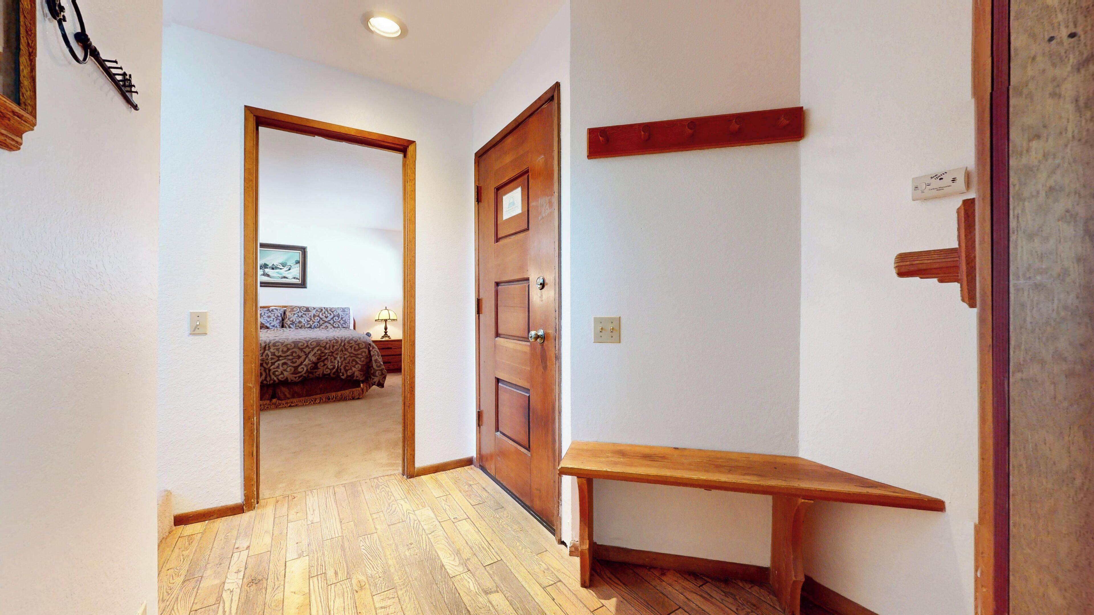 A hallway with a wooden bench, a wooden door, and a bed with patterned bedding in the background.
