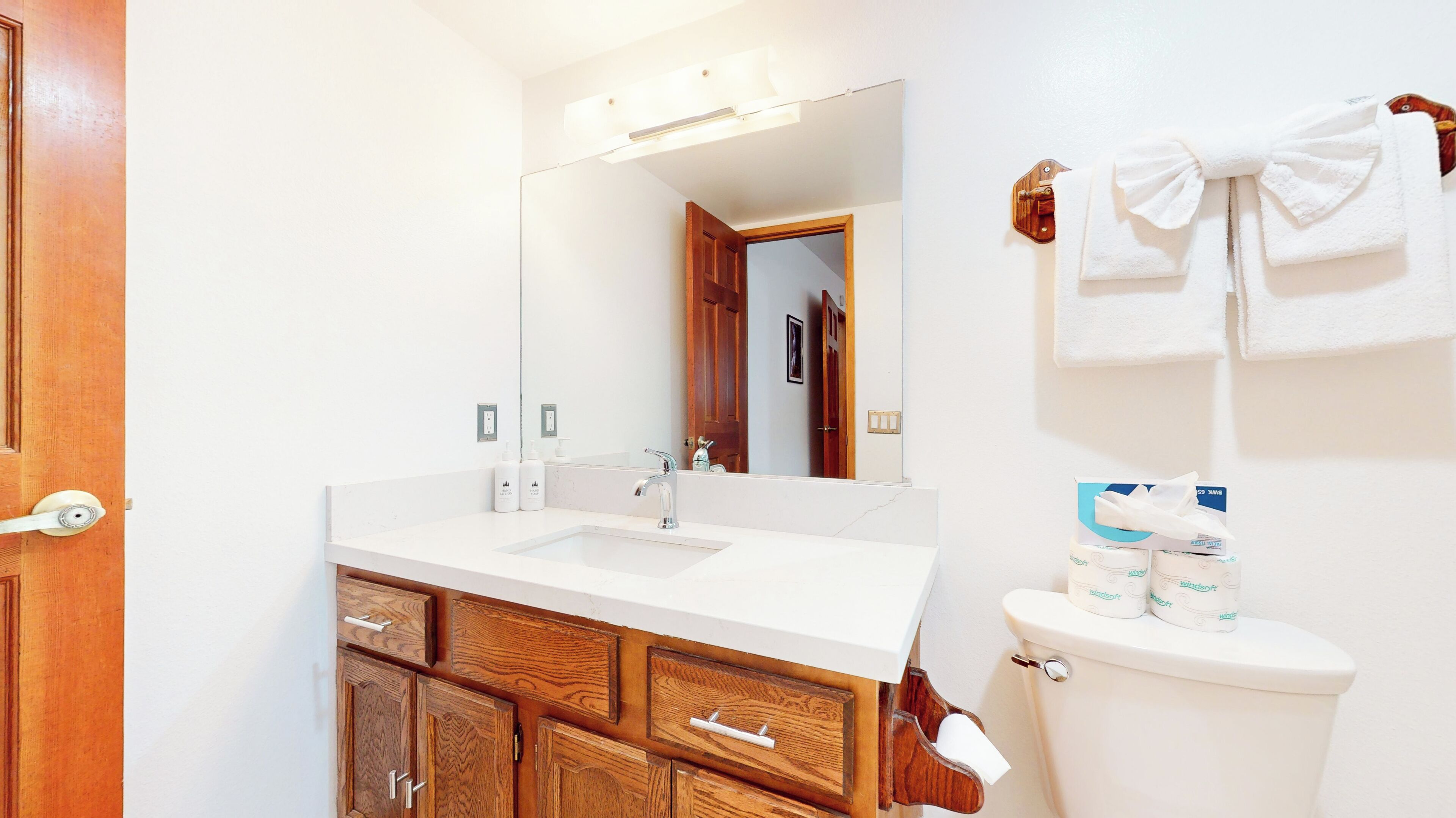 A bathroom with a white sink, wooden cabinets, a mirror, and a toilet with toilet paper and a towel rack.