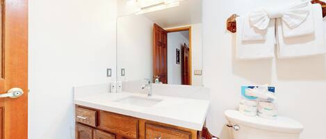 A bathroom with a white sink, wooden cabinets, a mirror, and a toilet with toilet paper and a towel rack.