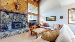 A cozy living room with a stone fireplace, wooden beams, a stone wall, a large window, a sofa, and a bear head decoration. - Rustic Home, Quiet Location. Near Bike Trails & Lakes Basin. Hot Tub On-site (Mammoth Lakes)