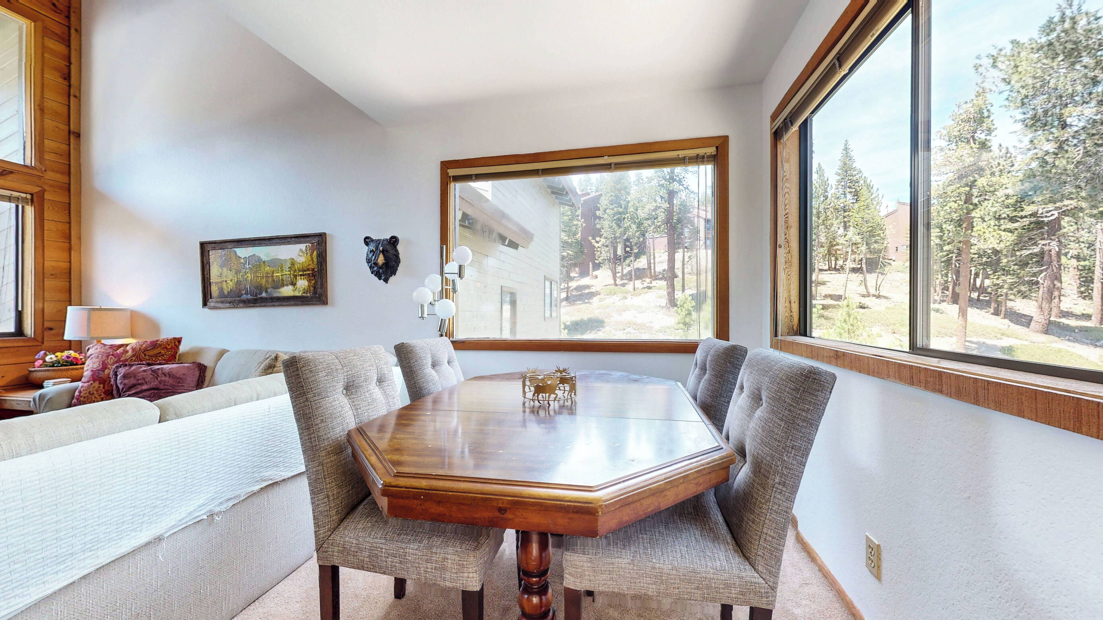 Une salle à manger avec une table en bois et des chaises rembourrées, une tête d’ours servant de décoration, un tableau et une fenêtre donnant sur des arbres.
