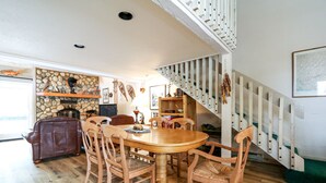 A dining area with a wooden table and chairs, a stone fireplace, and a staircase with white railings.
