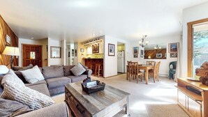 A living room with a sofa, coffee table, and a dining area with a table and chairs. There is a kitchen in the background with a refrigerator and a wooden wall.
