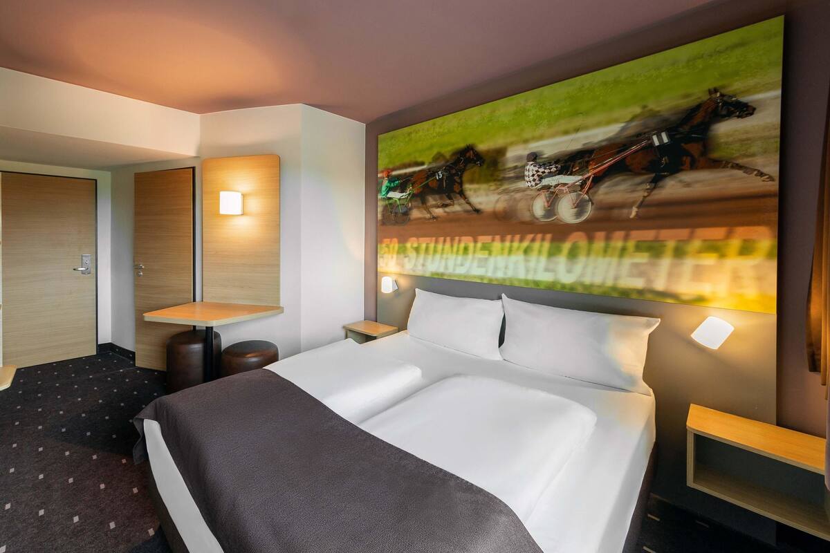 Ein modernes Hotelzimmer mit einem großen Bett, einem Schreibtisch und einem an der Wand befestigten Fernseher, der ein Pferderennen zeigt.