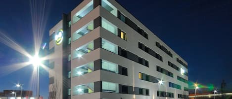 Un hotel de varios pisos con diseño moderno, que cuenta con balcones y un estacionamiento con varios coches.