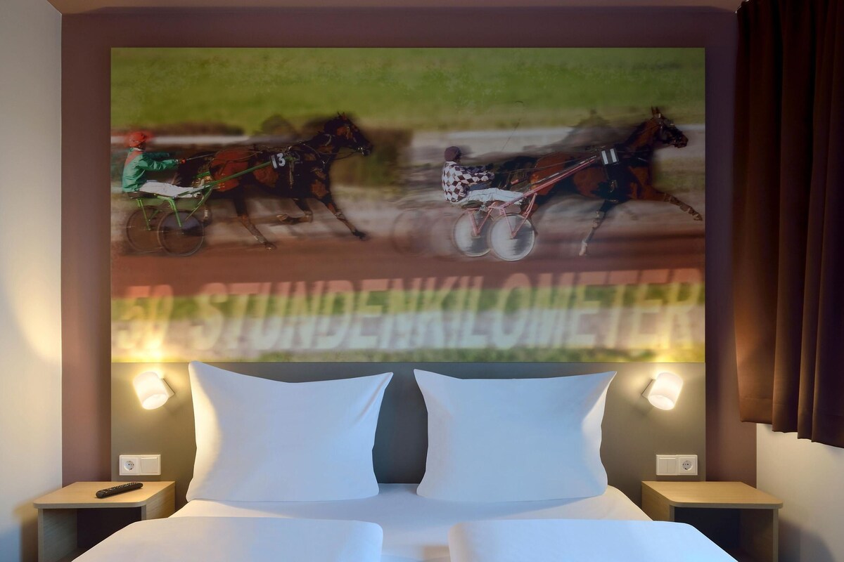 Ett hotellrum med en säng, två sängbord och ett väggmonterat konstverk som föreställer hästar i ett lopp.