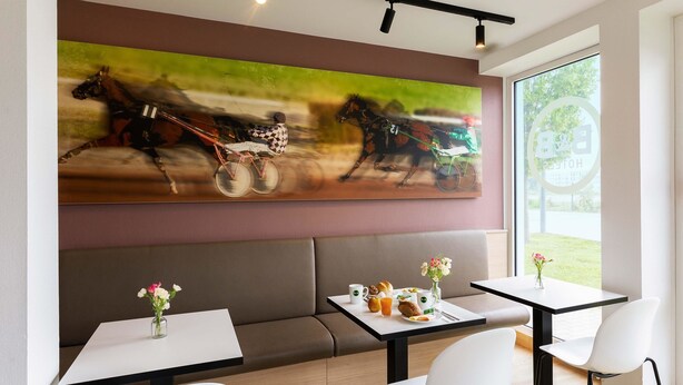 Un espace de repas moderne agrémenté d’un grand tableau représentant des chevaux, d’un canapé et de tables décorées de compositions florales.