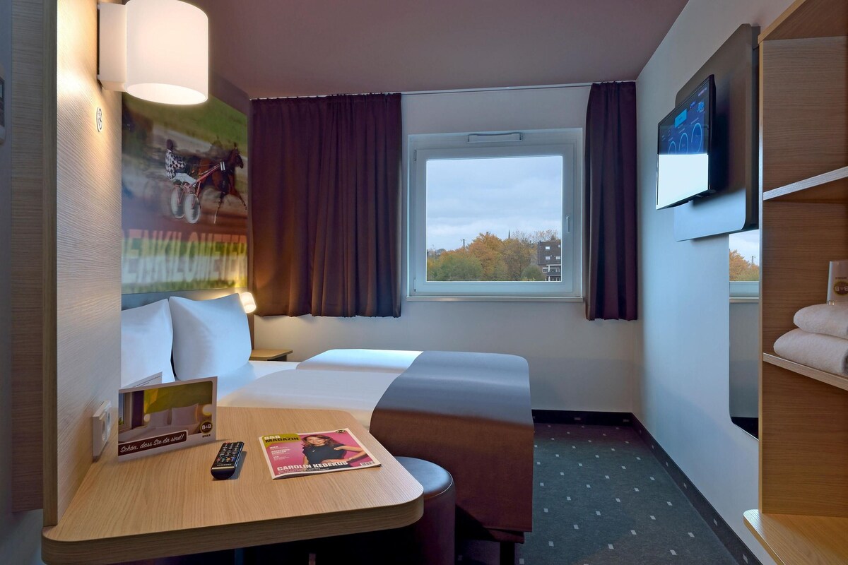 Ett hotellrum med en säng, ett skrivbord med en fjärrkontroll och en tidning, en tv på väggen och ett fönster med gardiner.